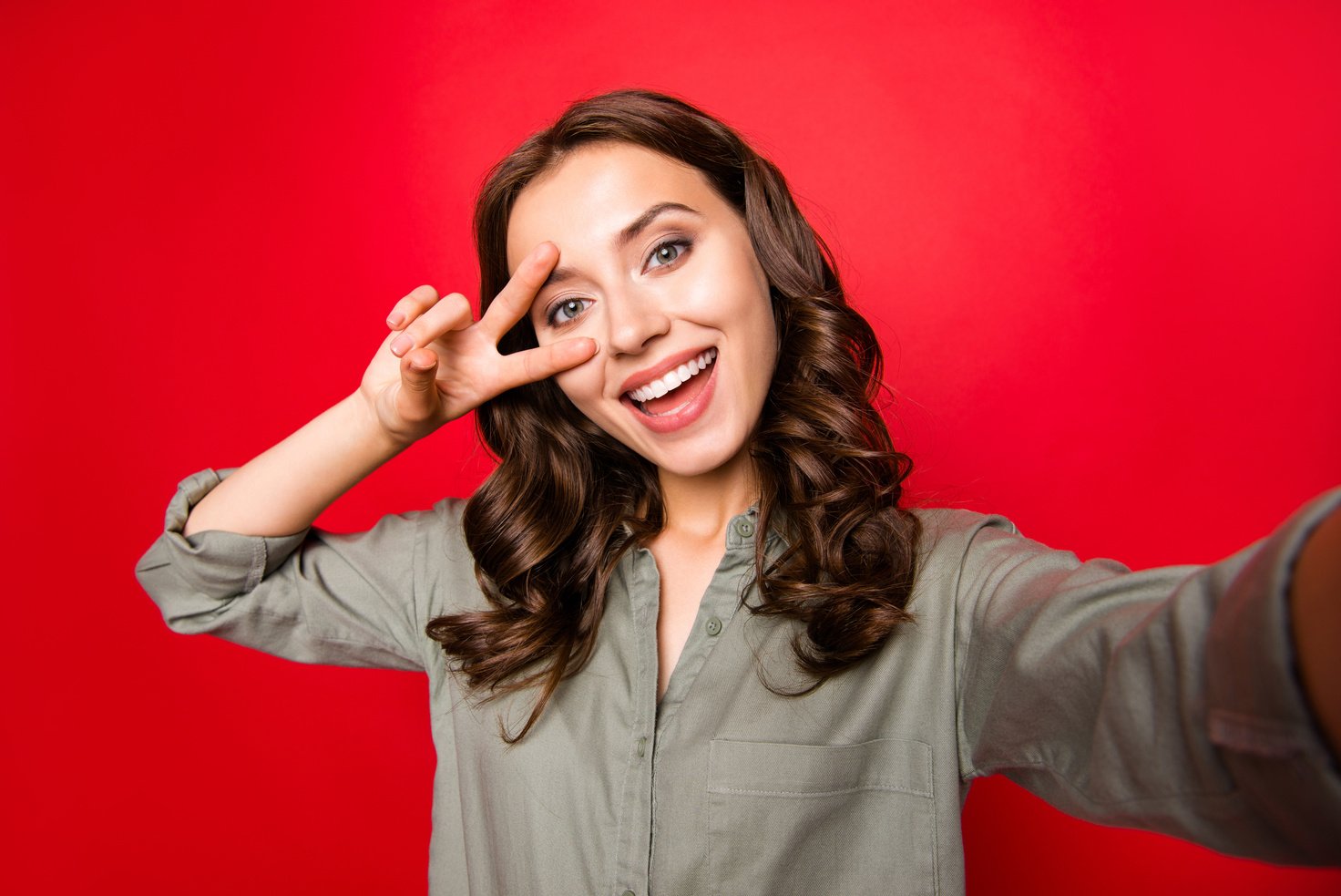 Stylish, good-looking lady with curly hairstyle, take selfie on front camera of modern smartphone, wear in casual blouse and make v-sign near eye isolated on shine red background